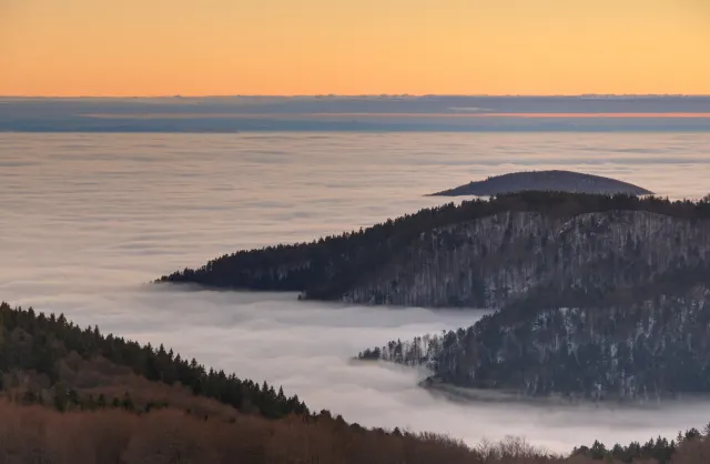 Dark forested ridges rise from a sea of fog under an orange sunset sky.