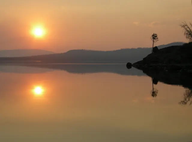 An orange sun and its reflection over a calm lake, with hazy hills and a lone tree on a rocky point in silhouette.