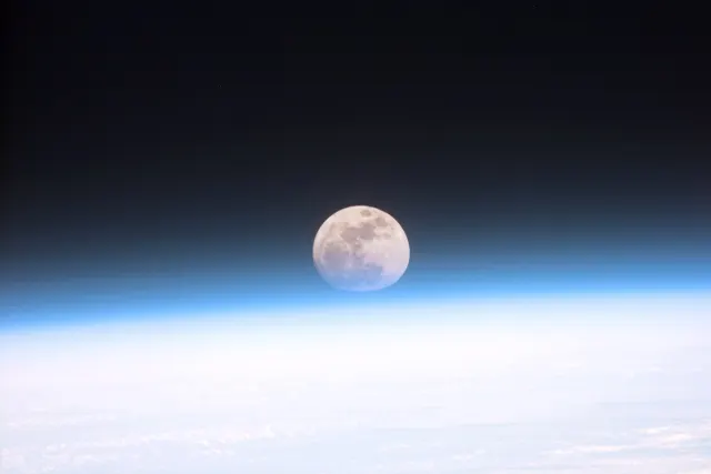 A pale full Moon above Earth's glowing blue and white atmospheric horizon against black space.
