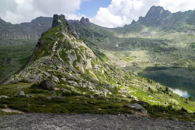 A pale rocky ridge rises beside a green valley and a dark mountain lake under a cloudy sky.