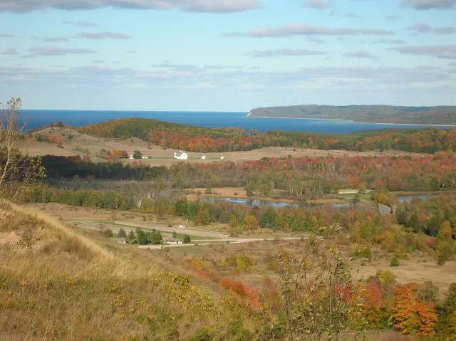 Autumn-colored woods and fields in a valley, with a white farm building and a blue lake along the horizon.