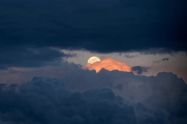 A pale full moon peeks through a gap in dark blue clouds above a glowing orange cloud just after sunset.