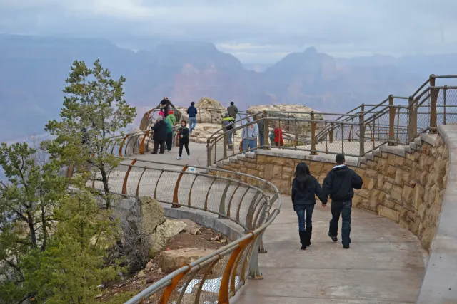 Two people walk hand in hand along a curved railed canyon-rim path, with hazy layered ridges beyond under an overcast sky.