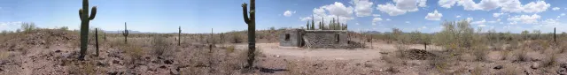 Wide desert panorama with two saguaro cacti and a small stone ranch house under a blue sky with light clouds.
