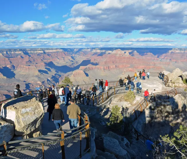 Visitors walk a railed path on a canyon rim above red rock buttes under a blue sky with large clouds.