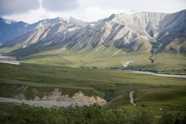 A dirt trail curves across a green tundra valley beneath pale, gullied mountains and a cloudy sky.