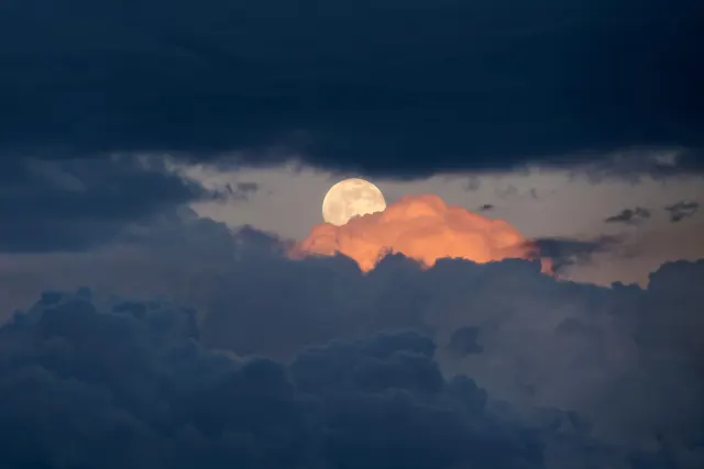 A pale full moon between dark blue storm clouds, with one orange-lit cloud glowing just below it at dusk.