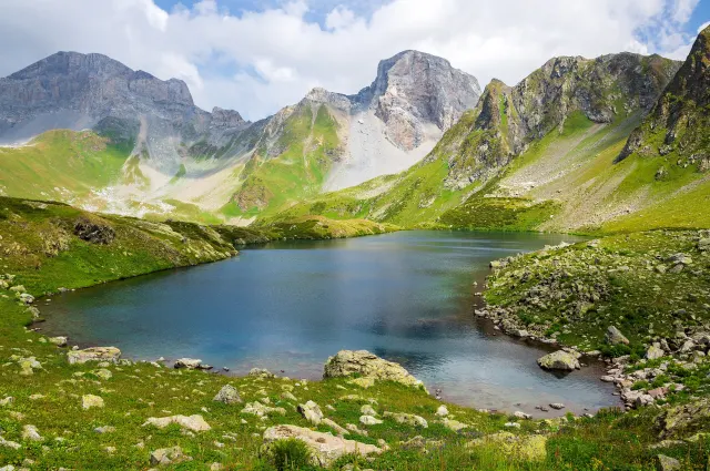 A blue-green mountain lake in a grassy cirque, with rocky peaks and scree slopes under a partly cloudy sky.