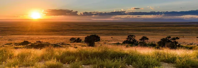 Orange sun setting over a wide grassy plain with dark shrubs and a band of clouds across the sky.