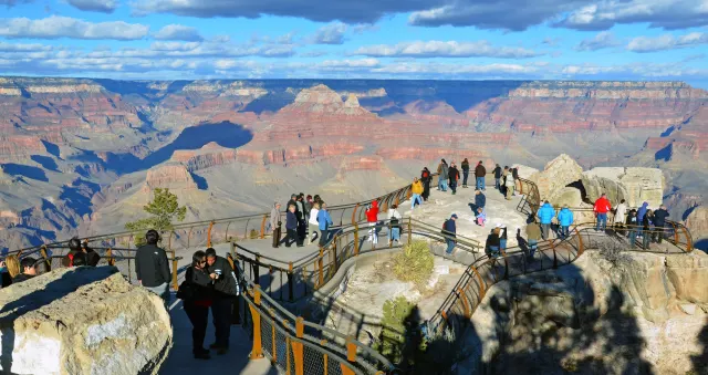 Visitors stand along railed overlook paths above a wide red-rock canyon with a large butte under a blue, partly cloudy sky.