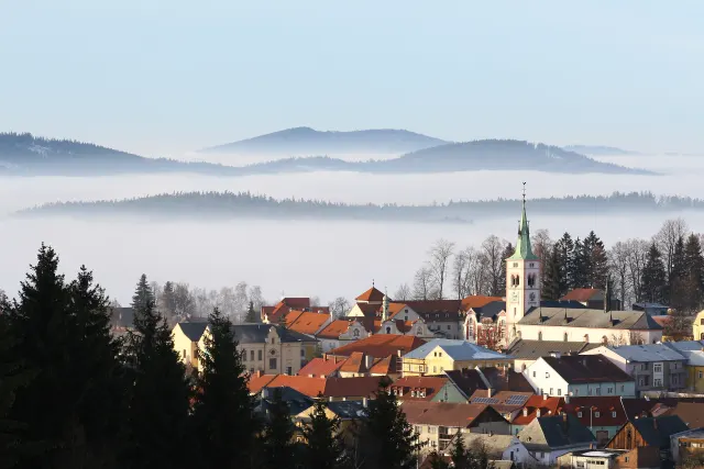 A town with a white church tower and red roofs beneath layered forested hills rising from a flat sea of fog.
