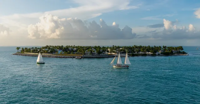 Two sailboats on turquoise water in front of a palm-covered island under large golden-lit clouds.